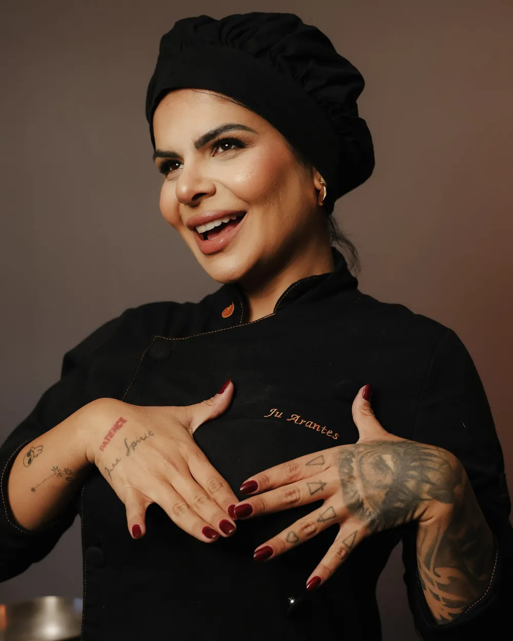 A chef Juliana Arantes de dólmã preta, sorrindo, com as mãos sobre o nome bordado.